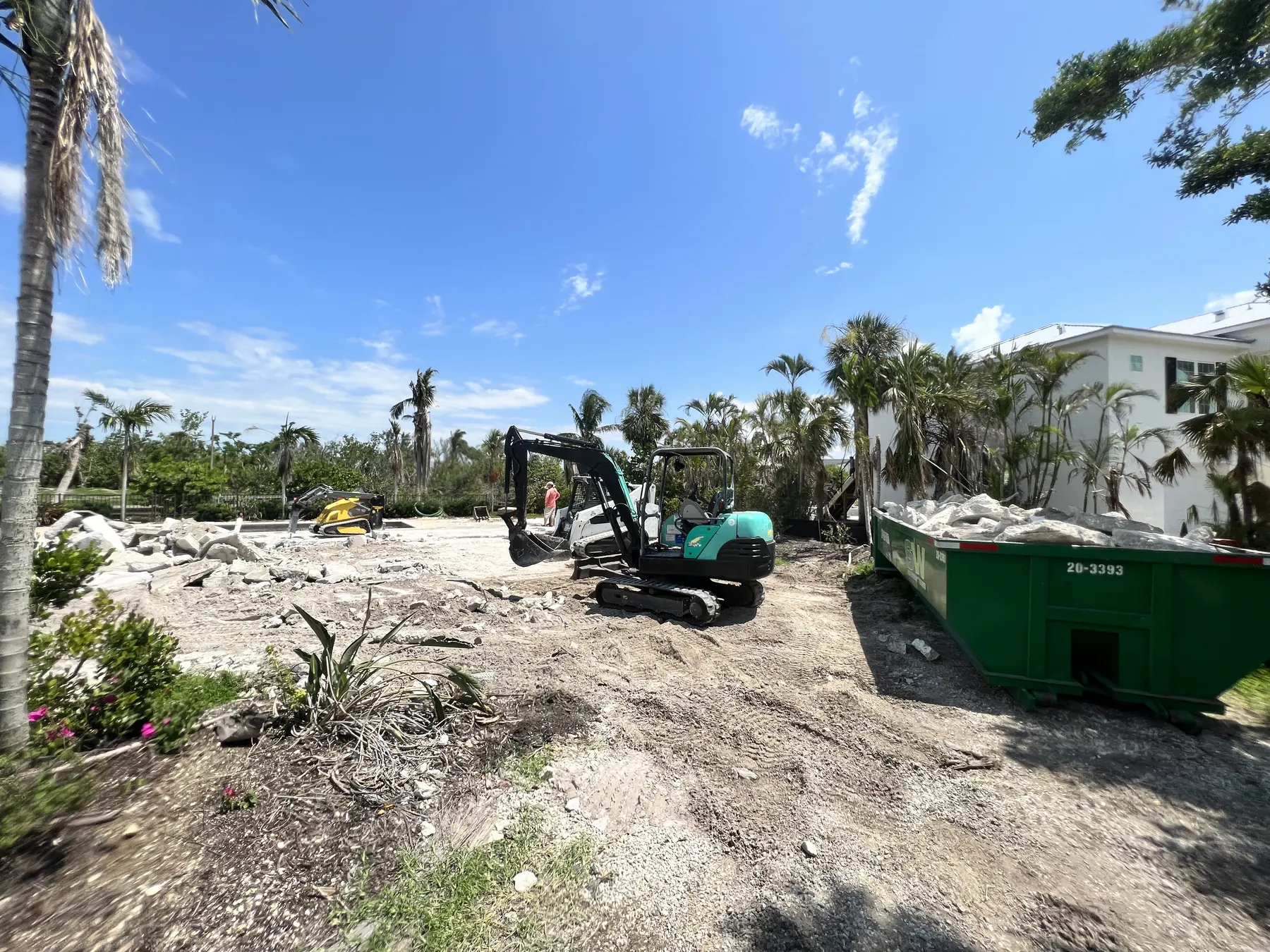 Excavator grading a Southwest Florida construction site with palm trees and a roll-off container nearby